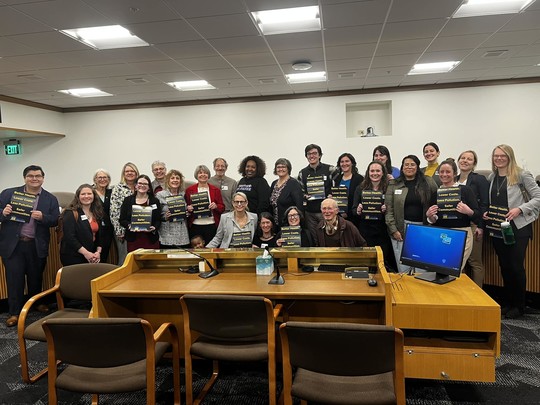A group of people with Senator Kate Lieber in the middle, holding signs that read "Better Health. Lower Costs. Less Pollution. Safer Communities"