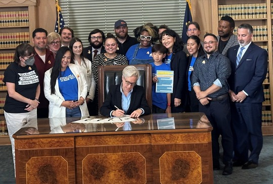 La gobernadora Kotek en su escritorio firmando un proyecto de ley con gente detrás de ella.