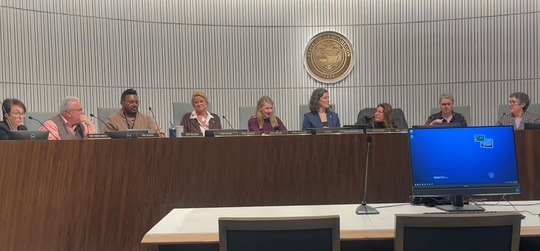 Rep Nelson at dias with other members of the committee with a light grey wall and state seal behind them