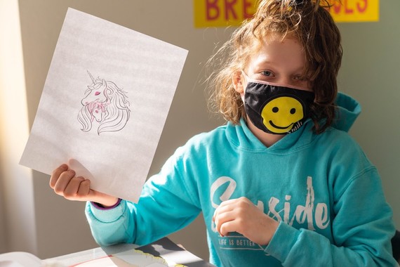Student in an aqua hoodie and a face mask with a smiley face holds up a drawing of a unicorn