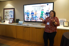 Kim Lyon speaks at the front of the room with a display screen behind her that shows a photo of kids on bikes