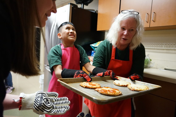 A middle school students pulls a tray with mini pizzas out of the oven as a teacher helps him