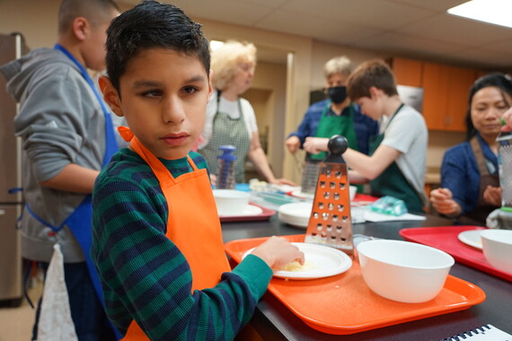 a student wearing an orange apron looks as the camera as he holds a piece of cheese he was shredding