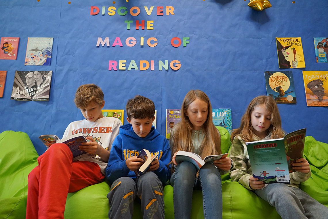 Elementary students read books on green bean bags in a library with a display that says discover the magic of reading