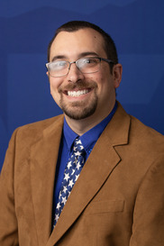 Smiling man with glasses, short dark hair, blue shirt, brown blazer, tie with stars