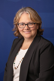 Smiling woman with blond hair, black rimmed glasses, black blazer, white shirt, beaded necklace