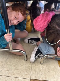 Girl with red hair, blue jacket, covers head and holds onto a table leg while practicing earthquake preparedness.