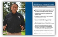 Native American man wearing blue Oxford shirt with FEMA logo, stands in front of trees, has hands extended to make a point