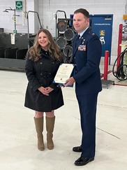 Woman on left in black coat, brown boots, wavy brown hair receives award from taller man in military dress uniform