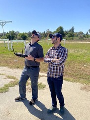 Two men in navy baseball caps stand in a field, one holds the controls to an unmanned aircraft system (UAS) 