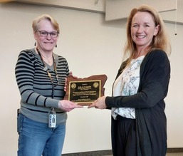Woman on left with short blonde hair, glasses, striped shirt receives plaque from woman on right with long strawberry blond hair