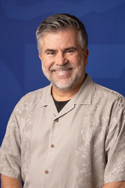 Smiling man, gray facial hair, wearing a tan Hawaiian shirt over a black T