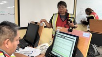 Tribal woman with short black hair, glasses and emermgency vest talks to a tribal man working on a computer