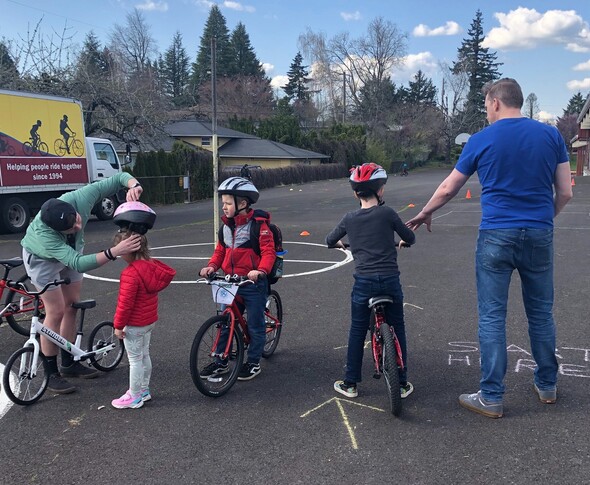 Three children practicing bicycling on a playground hard-top surface with the assistance of two adults on a sunny day.