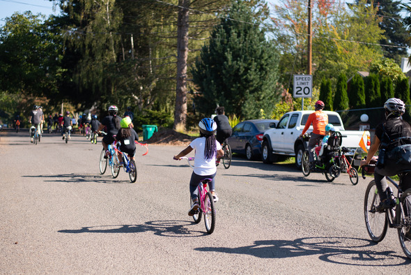 Dozens of bicyclists, young and old, riding down a 20 mph street on a sunny day. 
