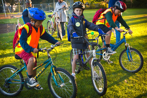 Three young boys wearing helmets and riding bikes on grass on a fall day.