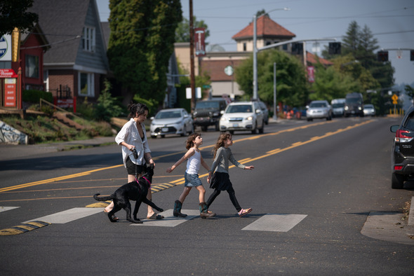 Two elementary school aged girls walk across a crosswalk with a woman and a black dog on a sunny day. 