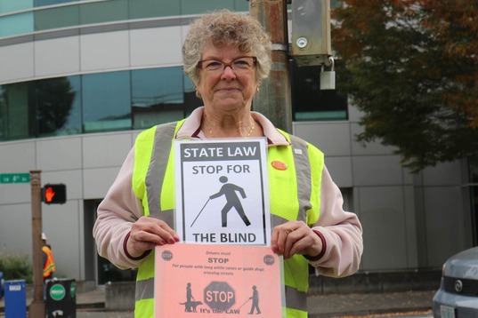 An older woman wearing a reflective safety vest holding a black and white 8.5x11 sign that reads "State law stop for the blind."