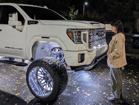 A large, white lifted GMC pickup truck's wheel are illuminated at night in a parking lot dwarfs an adult woman. 