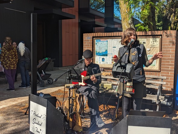 A woman on the right, standing, sings into a microphone while a man on the left, sitting, plays on guitar at St Johns Plaza