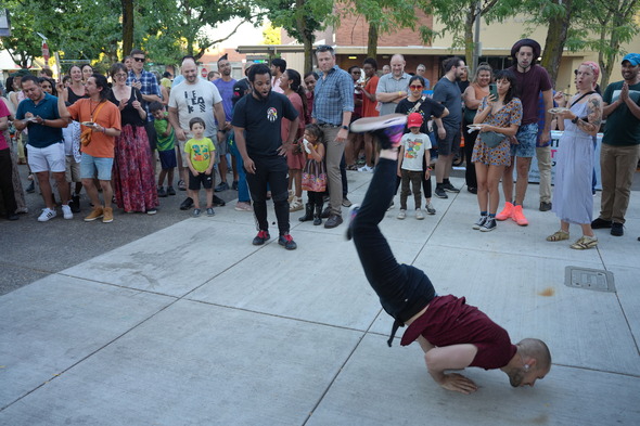 A large crowd of people in the background watch a man breakdancing in the foreground at St Johns Plaza