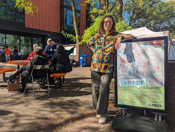 Tanya Harnett leans against the PBOT Public Plaza sign showing all the Plaza locations across Portland.
