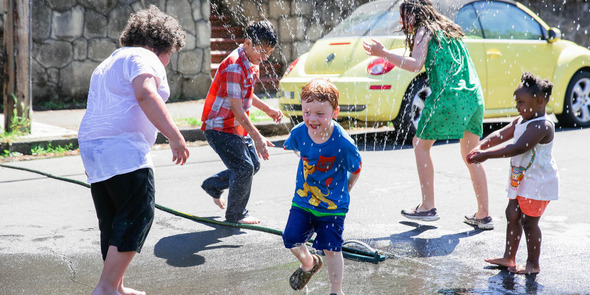 Five children of various ages playing around a water sprinkler on a street in the summer. 
