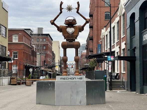 A robot stands in the middle of NW 13th Ave Plaza.  The robot has a large heart on its chest and holds a floating crystal between its hands