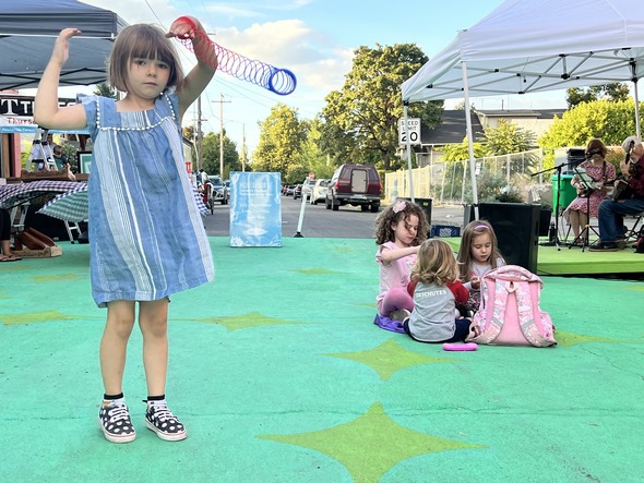 A child plays with a slinky while other children sit in a circle behind at Montavilla Plaza.  The street is painted green with yellow stars.
