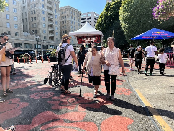 people are seen walking through Ankeny Alley plaza during a festival.  Two older women walk together.  One is using a cane.
