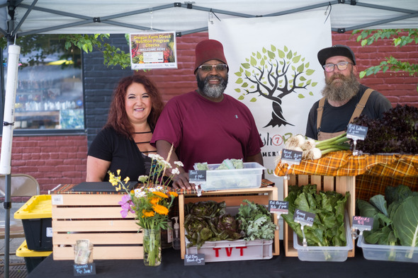 3 farmer's market vendors are smiling, standing behind their produce.