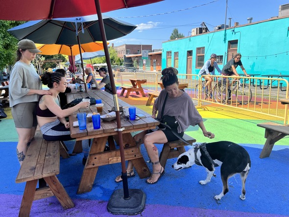 A group of people sit at a shaded picnic table with their dog at rainbow road.  The street is brightly painted with rainbows.
