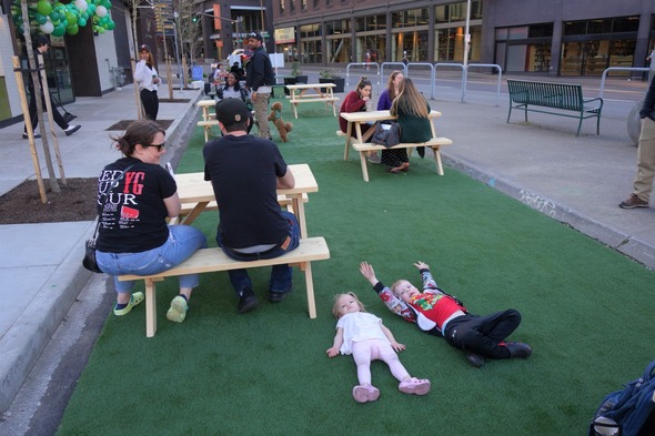 Two children lie on the astroturf while other guests of the plaza sit at a line of orange picnic tables at Pod Plaza.