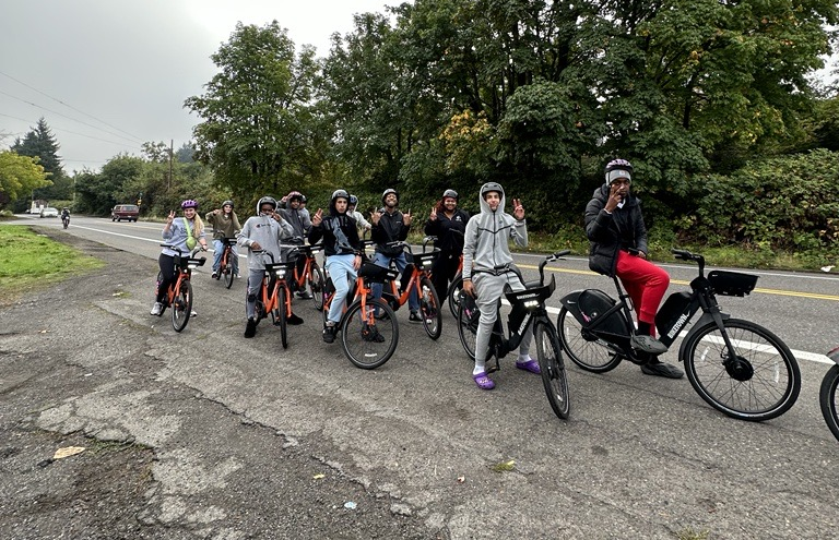 About a dozen high school students riding orange and black BIKETOWN bikes on a street next to a green area with shrubs, trees, and bushes.