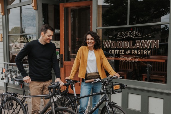 Jeri (wearing a yellow sweater) and a fellow adult standing behind their bikes on the sidewalk in front of the Woodlawn Coffee and Pastry storefront.