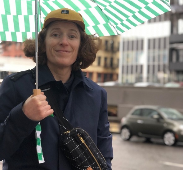 A person with short orange hair, wearing a yellow cap and blue jackets, and holding a green and white checkered umbrella on a city street..