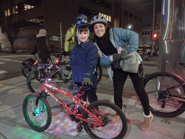 An adult and middle school-aged kid wearing winter gear and standing with their bikes wrapped in strings of lights at night. 