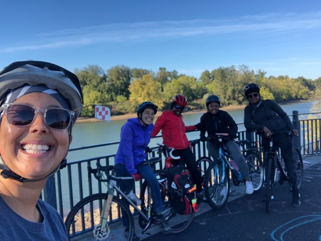 A group of five bicyclists taking a selfie photo next to a railing next to the a river waterfront with heavy foliage on the opposite bank.