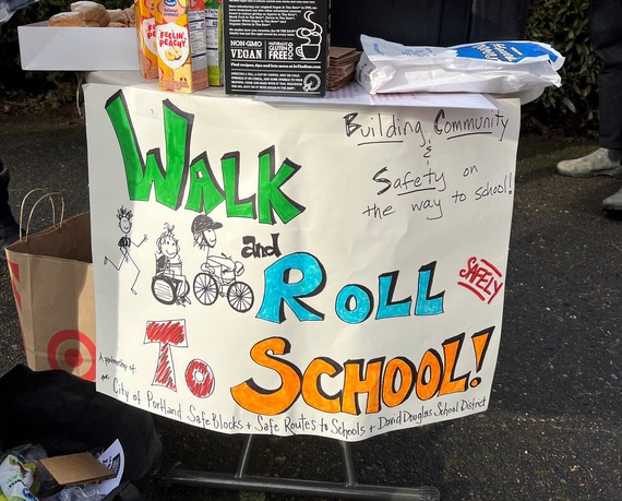 A colorful, homemade "walk and roll to school" sign attached to the front of a pop-up snack table outside on a sidewalk.