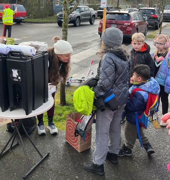 A group of four elementary school-aged students huddle around a sidewalk pop-up where two adults are serving hot beverages.