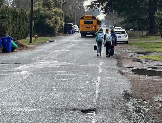 Three middle school students with backpacks on walk on a street without sidewalks behind a school bus on an overcast rainy day. 