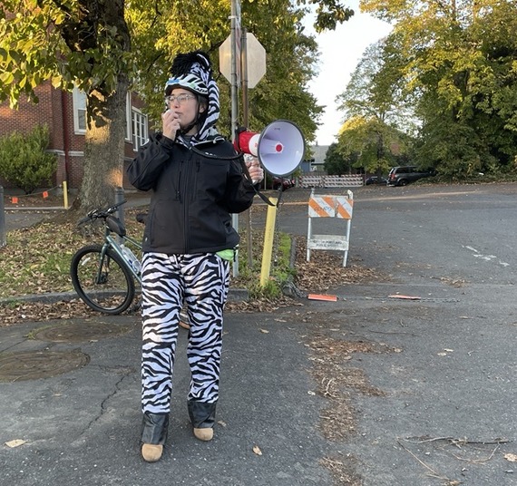 An adult wearing a zebra costume under a black jacket speaking into a megaphone on an autumnal street.