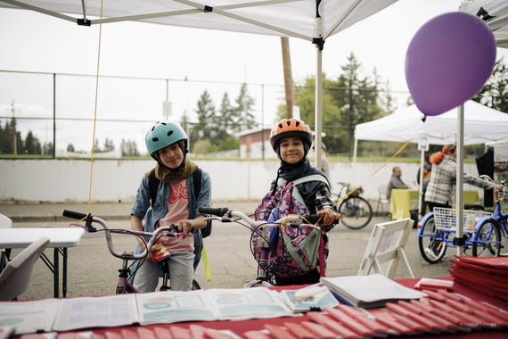 Two elementary school-aged youth on bikes wearing helmets stand in front of a table at a community event.