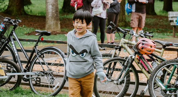 Two elementary school-aged youth ride on the back of a cargo bike that an adult is pedaling on a neighborhood street.