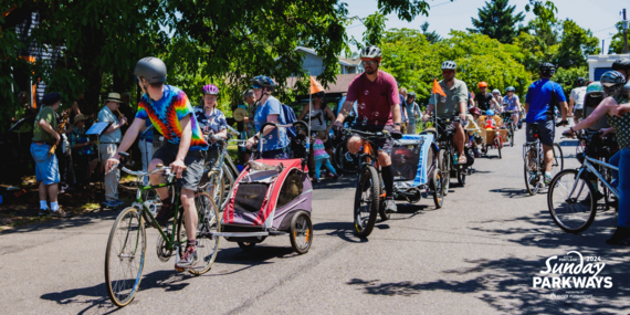 Dozens of people wearing bright, colorful clothes and riding bikes on a neighborhood street on a sunny day.