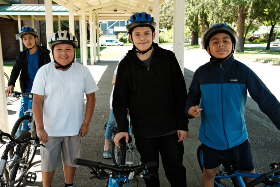 Middle school students wearing helmets and standing with their bikes under a covered awning.