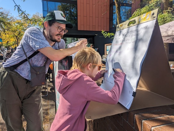 An adult watches as a young child writes their response to the question, "what events do you want to see at public plazas?"