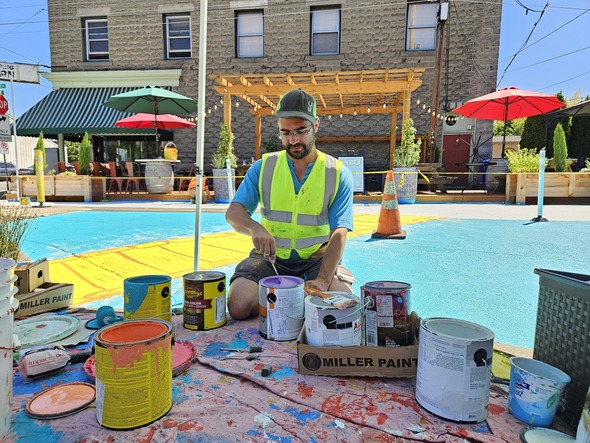 A man in a yellow vest kneels in front of a tarp of paint supplies. Behind him is a colorfully painted plaza with seating.