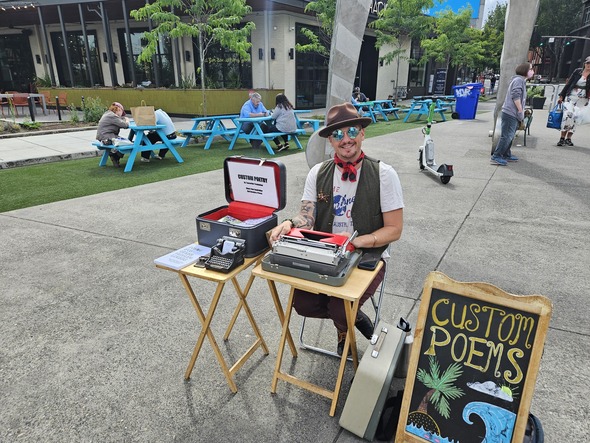 A man sits at a small table at pod plaza offering custom poems.