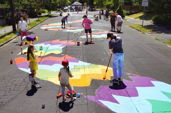 A group of young and old paint a long stretch of street with brushes and rollers. The painting is of a long serpent-like friendly dragon.